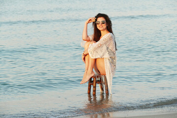 A young beautiful woman poses on the ocean beach. Pretty brunette in white beach tunic and black...