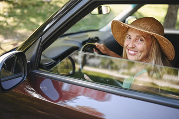 Fototapeta premium Happy senior woman driver in straw hat driving sitting in new car, smiling looking at camera enjoying journey. Driving courses and life insurance. Retired people activity and road trip concept 