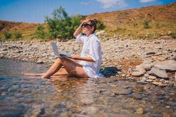 Young woman freelancer traveler wearing white shirt anywhere working online outdoors using laptop enjoying lake view. Happy female downshifter in sunglasses holding computer at sea coastline 