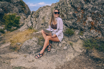 Young woman freelancer traveler working online anywhere outdoors using laptop enjoying mountain peak view. Happy female downshifter in sunglasses holding computer in unusual workplace at summer