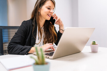 Young brunette executive girl in American jacket scrolling on her laptop while talking on the phone. Entrepreneur woman.