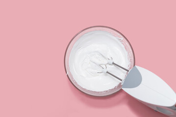 Bowl with whipped egg whites and mixer on pink table background, top view
