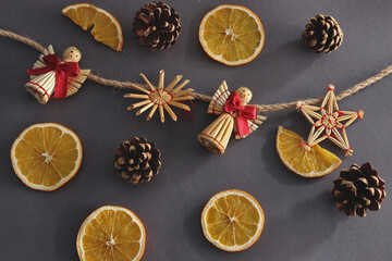 straw ornaments in the shape of an angel, stars and snowflakes on a string, cut slices of dried orange, cones on brown background