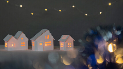 Small white cardboard houses with cut shining windows on dark blue bokeh blurred foreground and background. Shallow depth of focus. Winter decoration set with sparkling lights, handmade folded models.