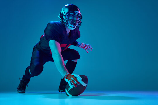 Sportive, strong man, american football player in sports team uniform and protective helmet isolated over blue background in neon light. Power, energy, achievements, skills
