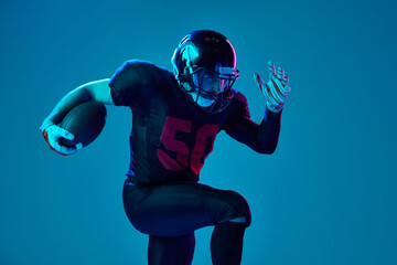Sportive, strong man, american football player in sports team uniform and protective helmet isolated over blue background in neon light. Power, energy, achievements, skills