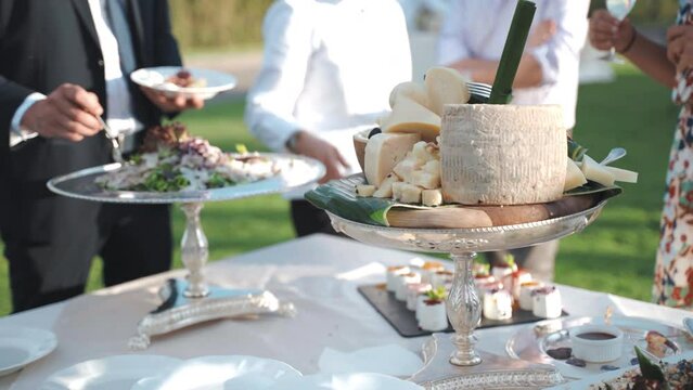Luxury banquet tables on open-air terrace, waiter in uniform serving invited guest, variety of food delicacy on silver trays. Fresh lettuce, parmigiano cheese, nuts and other appetizers and snacks for