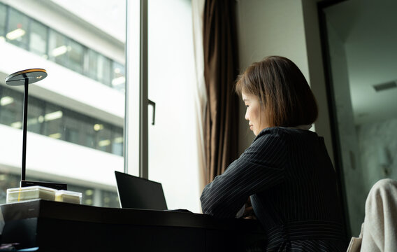 A young woman is seated at a desk and typing on a computer. Asian businesswoman in front of window