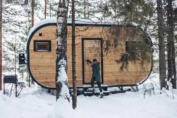 Girl walking in winter snowy forest near wooden tiny house. Winter landscape. Glamping