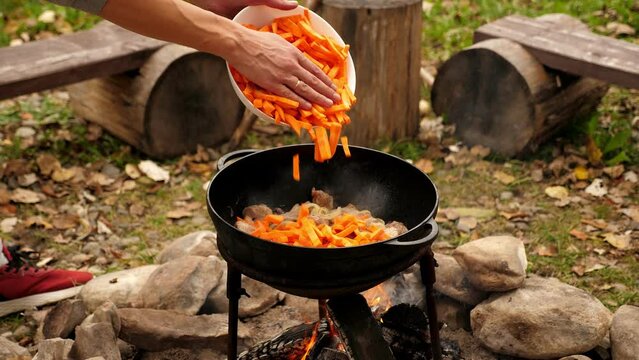 Close-up of a man cooking pilaf in a cauldron on a fire, he puts carrots. Cooking in an open oven. Cooking food in nature on the cauldron.