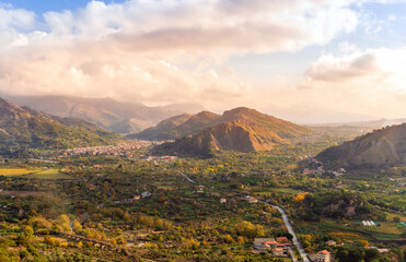 beautiful landscape with amazing view from highland with green slopes and bushes to a valley with town, road and nice mountains and scenic cloudy sunset on background