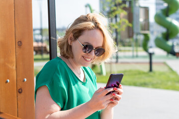 Portrait of a smiling woman in sunglasses, holding a smartphone in her hands, sitting on a bench in a city park.