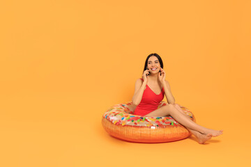 Happy young woman with beautiful suntan talking by phone on inflatable ring against orange background, space for text