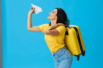 Travel woman with yellow suitcase, passport and ticket in hand, paper plane, in yellow T-shirt on blue background, happiness from travel, glasses, copy space