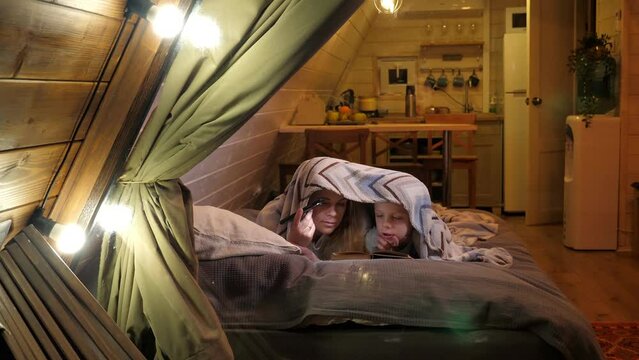 A mother and son are lying under a blanket with a flashlight and reading a book in a wooden triangular country house, a view through a window with flashlights and a thick curtain.