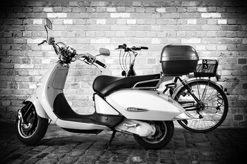 Black and white Picture of a Motor scooter in front of a bicycle on granite natural stone path with a brick wall as background