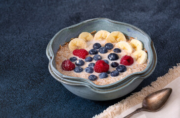 Plant-based bowl of steel cut oats with banana, blueberries, raspberries in a blue bowl on a dark blue surface with copy space
