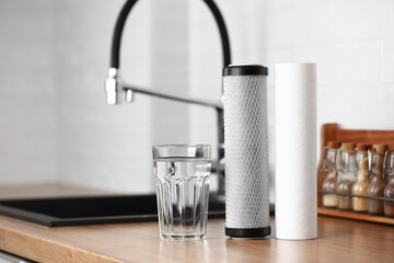 A glass of clean water and foamed polypropylene filter cartridges on wooden table in a kitchen interior. Installation of reverse osmosis water purification system. Concept Household filtration system.