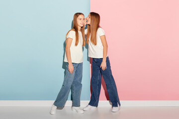 Two beautiful teen girls, siblings in t-shirts and jeans posing isolated over duotone pink-blue background. Emotions, family, fun, joy, leisure activities concept