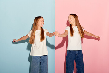 Happy beautiful teen girls, siblings in t-shirts and jeans dancing isolated over duotone pink-blue background. Emotions, family, fun, joy, leisure activities concept