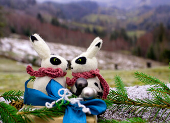 A pair of white and black felted rabbits surrounded by sprigs of a Christmas tree and cones against the background of a New Year's gift and a mountain landscape