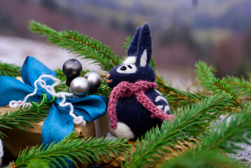A dark gray felted rabbit surrounded by sprigs of a Christmas tree and cones near a New Year's gift and against the backdrop of a mountain landscape