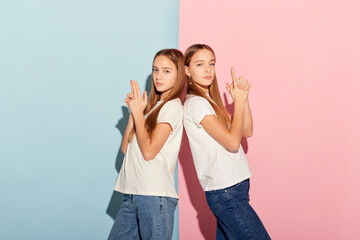 Adventure. Two beautiful teen girls, siblings in t-shirts and jeans looking at camera isolated over pink-blue background. Emotions, fun, joy, leisure activities concept