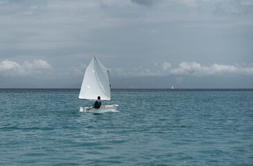 Obraz premium Lonely sailor on training sailing pram optimist education boat in the sea in Greece, water background and cloudy sky