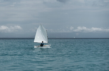 Lonely sailor on training sailing pram optimist education boat in the sea in Greece, water background and cloudy sky