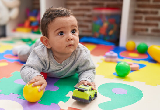 Adorable hispanic baby playing with ball and car lying on floor at kindergarten