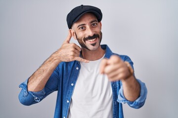 Hispanic man with beard standing over isolated background smiling doing talking on the telephone gesture and pointing to you. call me.