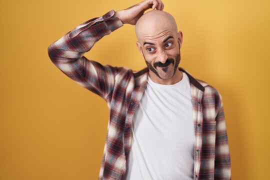 Hispanic man with beard standing over yellow background confuse and wonder about question. uncertain with doubt, thinking with hand on head. pensive concept.