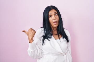 Mature hispanic woman standing over pink background surprised pointing with hand finger to the side, open mouth amazed expression.