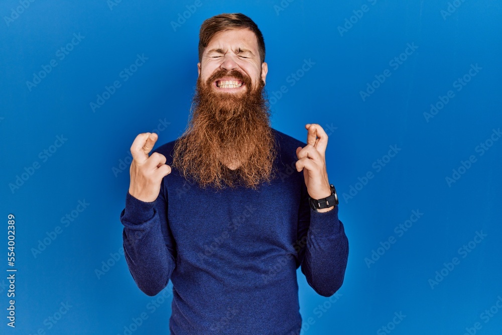 Canvas Prints Redhead man with long beard wearing casual blue sweater over blue background gesturing finger crossed smiling with hope and eyes closed. luck and superstitious concept.