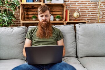 Redhead man with long beard using laptop sitting on the sofa at the living room thinking attitude...
