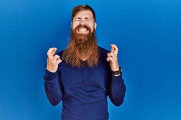 Redhead man with long beard wearing casual blue sweater over blue background gesturing finger crossed smiling with hope and eyes closed. luck and superstitious concept.