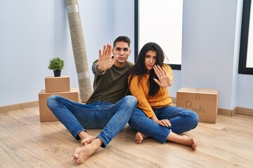 Fototapeta premium Young couple sitting on the floor at new home doing stop sing with palm of the hand. warning expression with negative and serious gesture on the face.