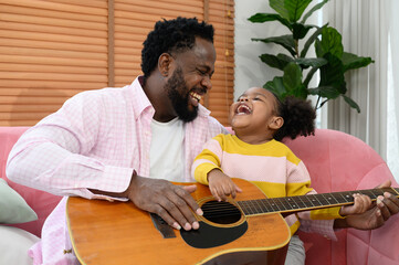 Fototapeta premium African girl and father are playing guitar while sitting on the sofa at home