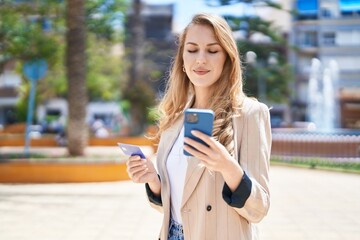 Young blonde woman using smartphone and credit card at park