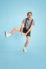 Fototapeta na wymiar Portrait of young man in checkered shirt, shorts and suspenders cheerfully jumping isolated over blue background. Success