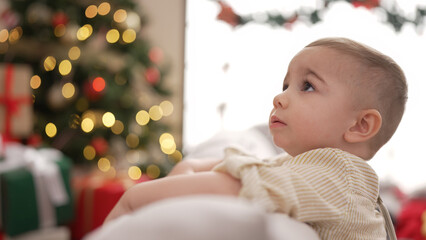 Adorable toddler sitting on sofa by christmas tree with serious expression at home