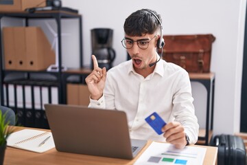 Young hispanic man working using computer laptop holding credit card surprised pointing with finger to the side, open mouth amazed expression.