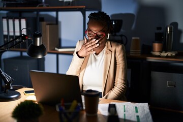 Beautiful black woman working at the office at night smelling something stinky and disgusting, intolerable smell, holding breath with fingers on nose. bad smell - obrazy, fototapety, plakaty