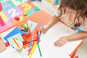 Adorable hispanic girl student drawing on paper sitting on table at kindergarten