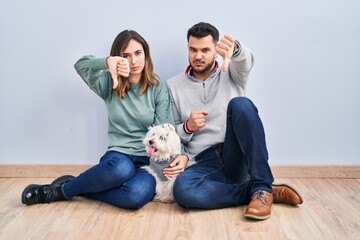 Young hispanic couple sitting on the floor with dog looking unhappy and angry showing rejection and negative with thumbs down gesture. bad expression.