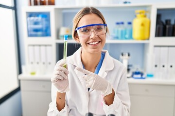 Young blonde woman working at scientist laboratory holding sample smiling happy pointing with hand and finger to the side