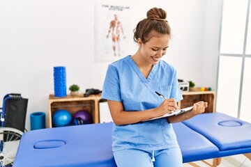 Young beautiful hispanic woman physiotherapist smiling confident writing on document at rehab clinic