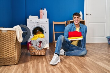 Young hispanic man holding clean laundry sticking tongue out happy with funny expression.