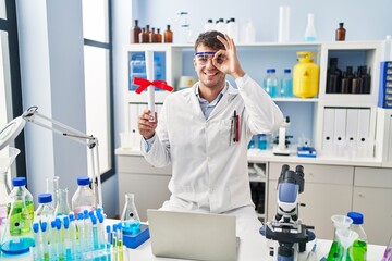 Young hispanic man working at scientist laboratory holding diploma smiling happy doing ok sign with hand on eye looking through fingers