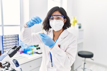 Young latin woman wearing scientist uniform and medical mask using pipette at laboratory